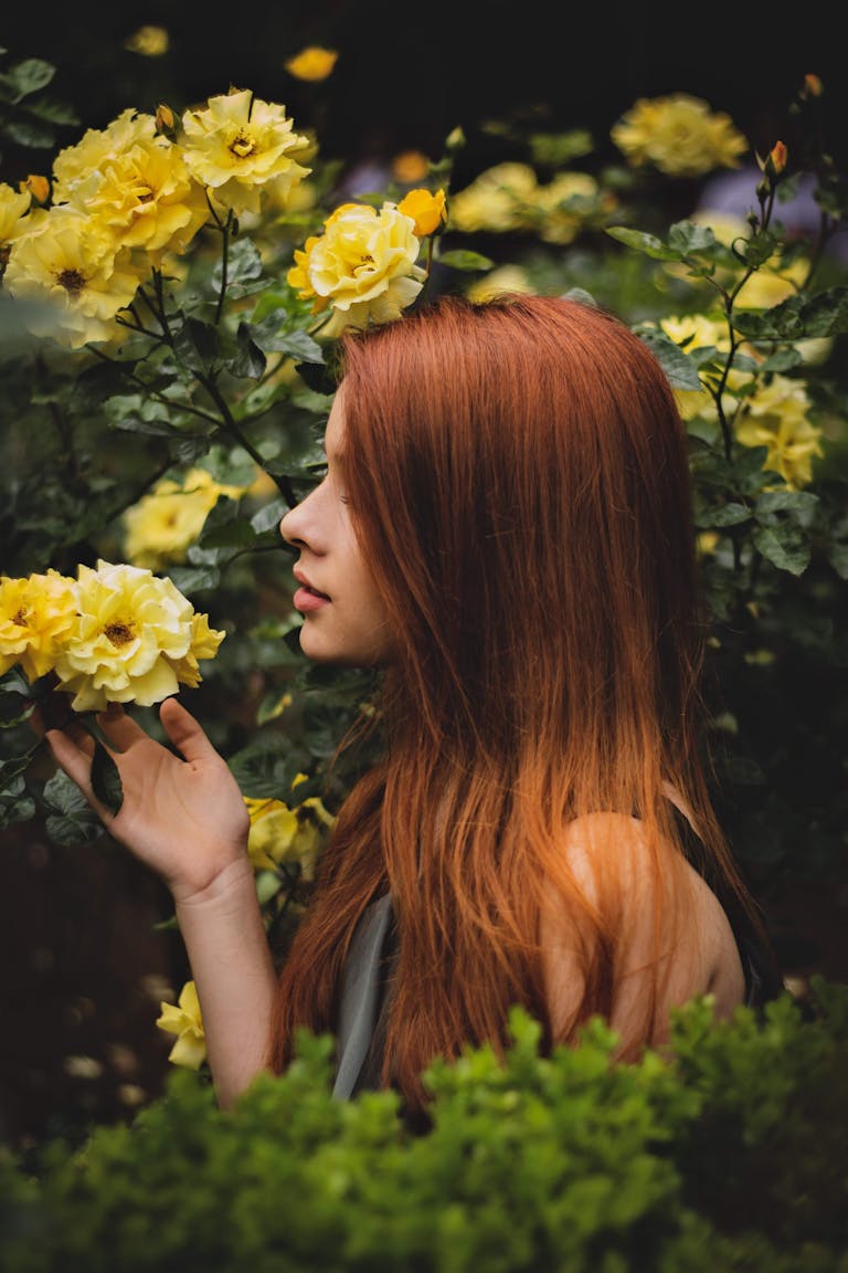 A woman with red hair enjoying the scent of blooming yellow roses in a garden.